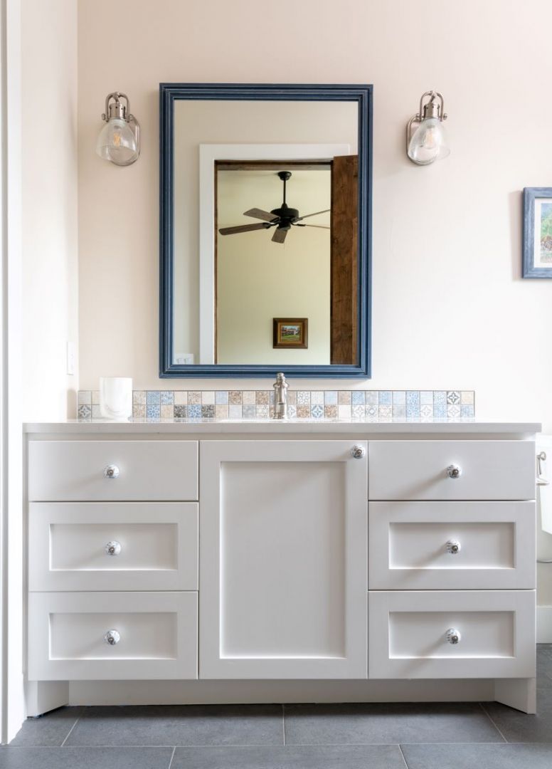 White bathroom vanity with blue-framed mirror, sconces, and patterned backsplash.