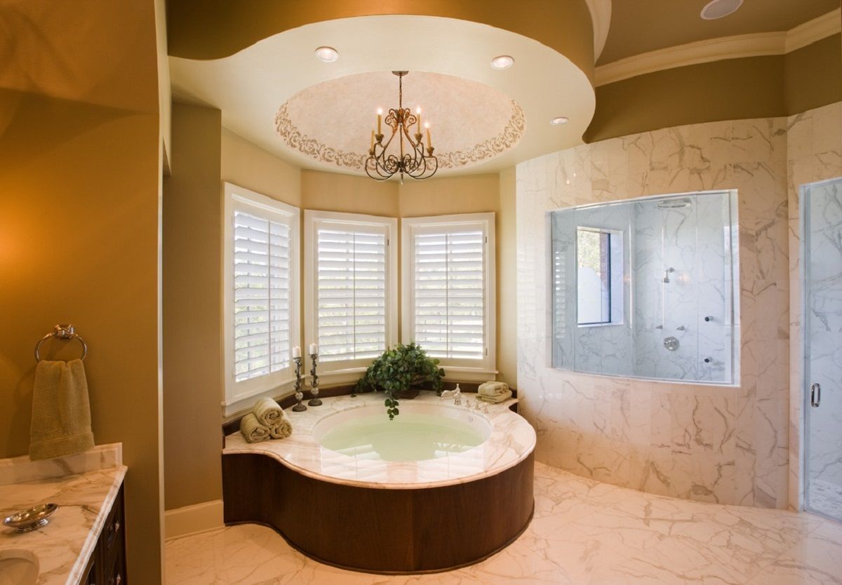 Luxury bathroom with a tub in front of shuttered windows, marble walls, and a chandelier.
