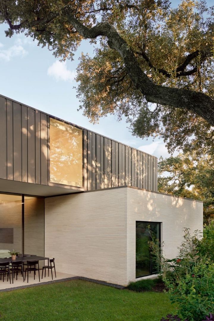 Modern two-story house with stone facade, metal siding, large windows, and a large oak tree in the yard.