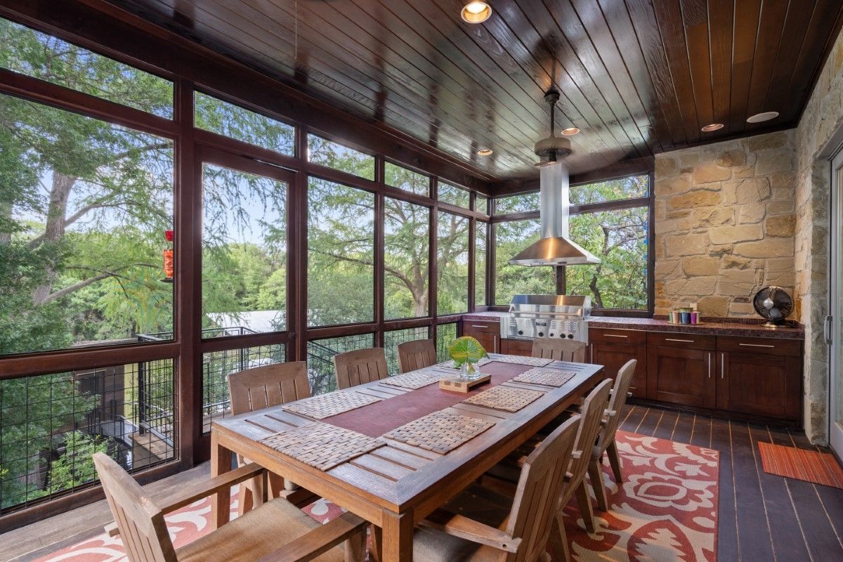 Screened-in dining porch with wooden table, chairs, and a view of trees through large windows.