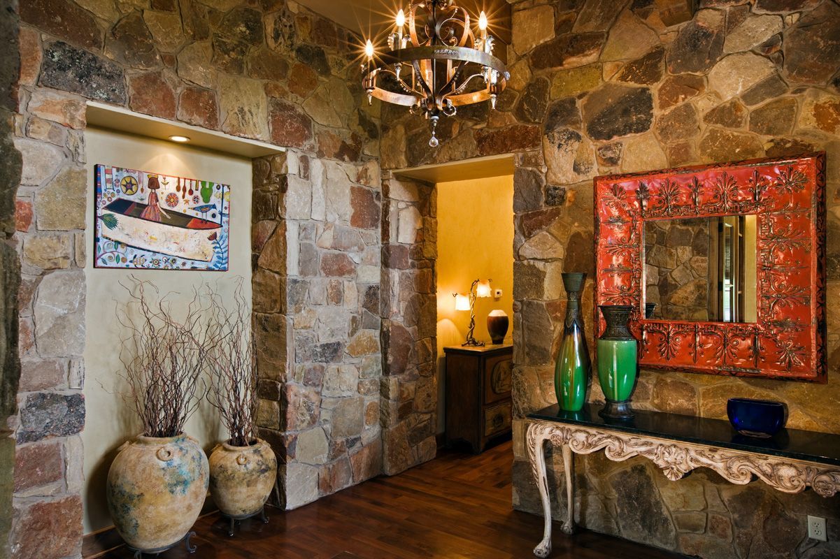 Stone-walled hallway with artwork, vases, and ornate table with a mirror and chandelier.