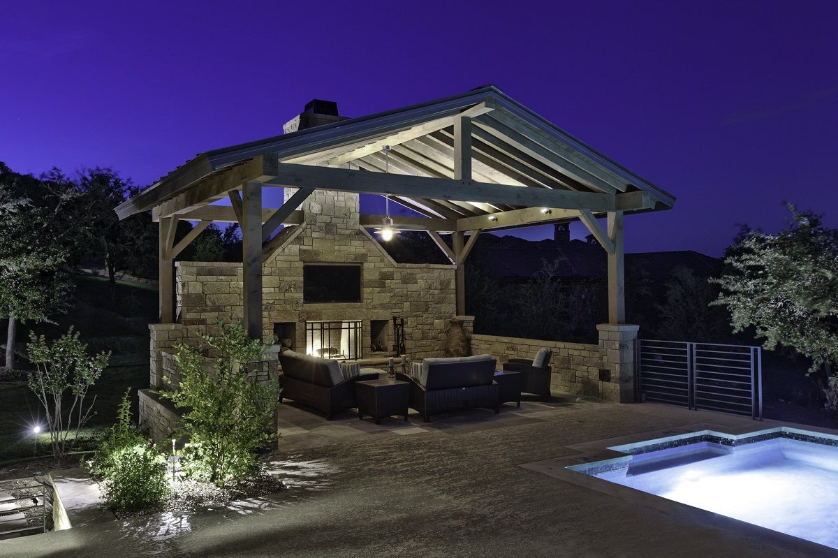 Outdoor stone fireplace with seating under a wooden roof, next to a lit swimming pool at night.