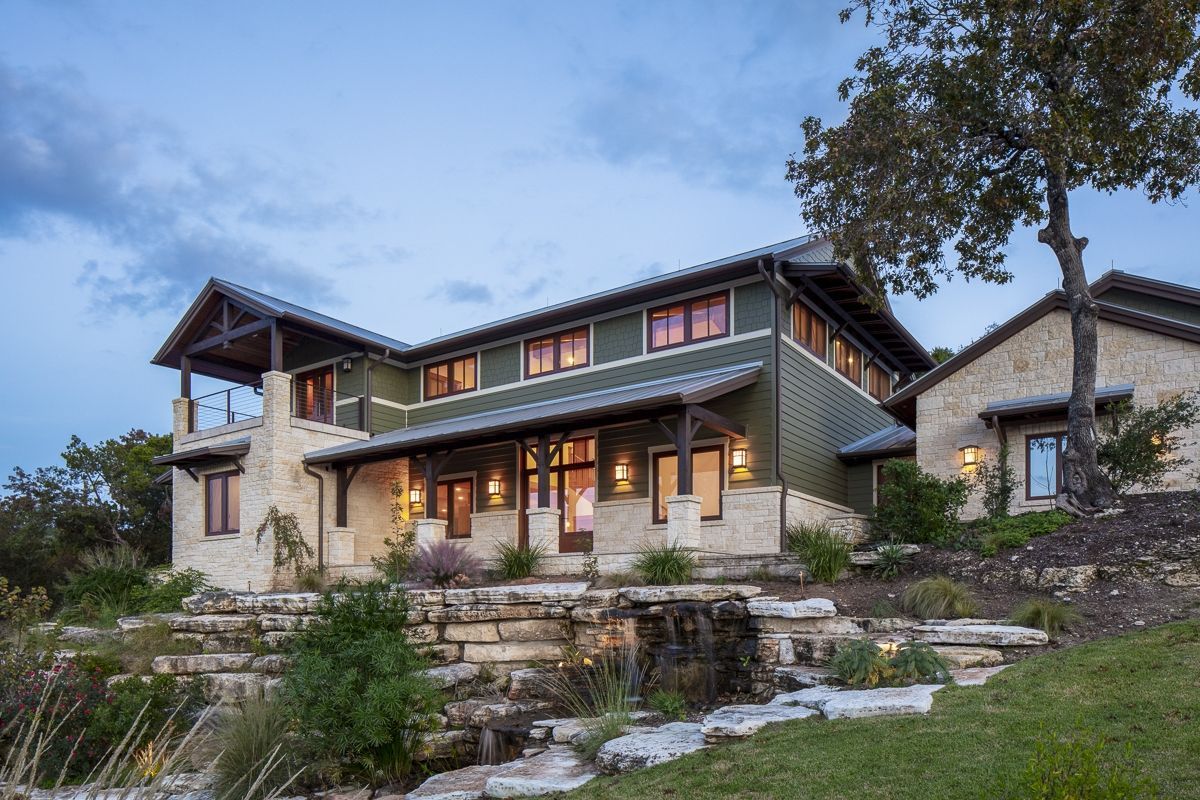 Two-story green house with stone accents, porch, and tree in a natural setting at dusk.