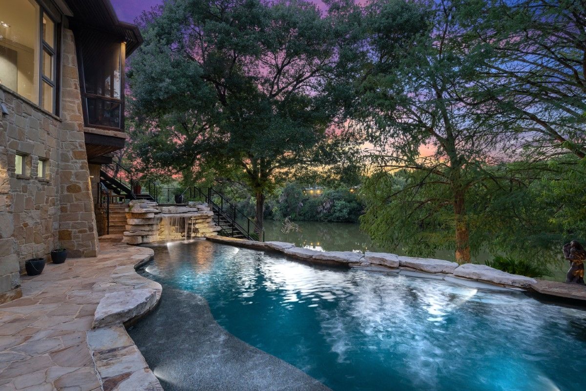 Infinity pool overlooking a body of water at dusk, stone patio, trees, and a multi-story stone building.