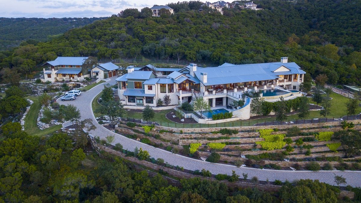 Aerial view of a large estate with light-colored buildings, pools, and a winding driveway, surrounded by trees.