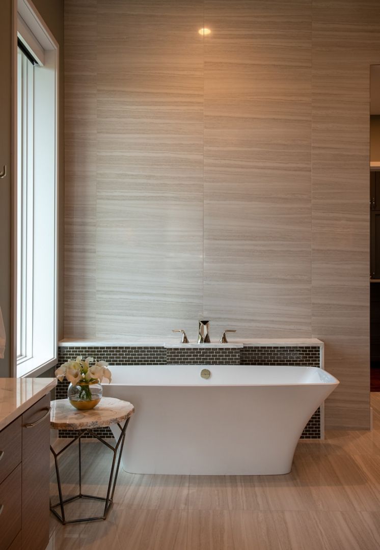 Modern bathroom with a white tub, beige tile wall, and small side table with flowers.