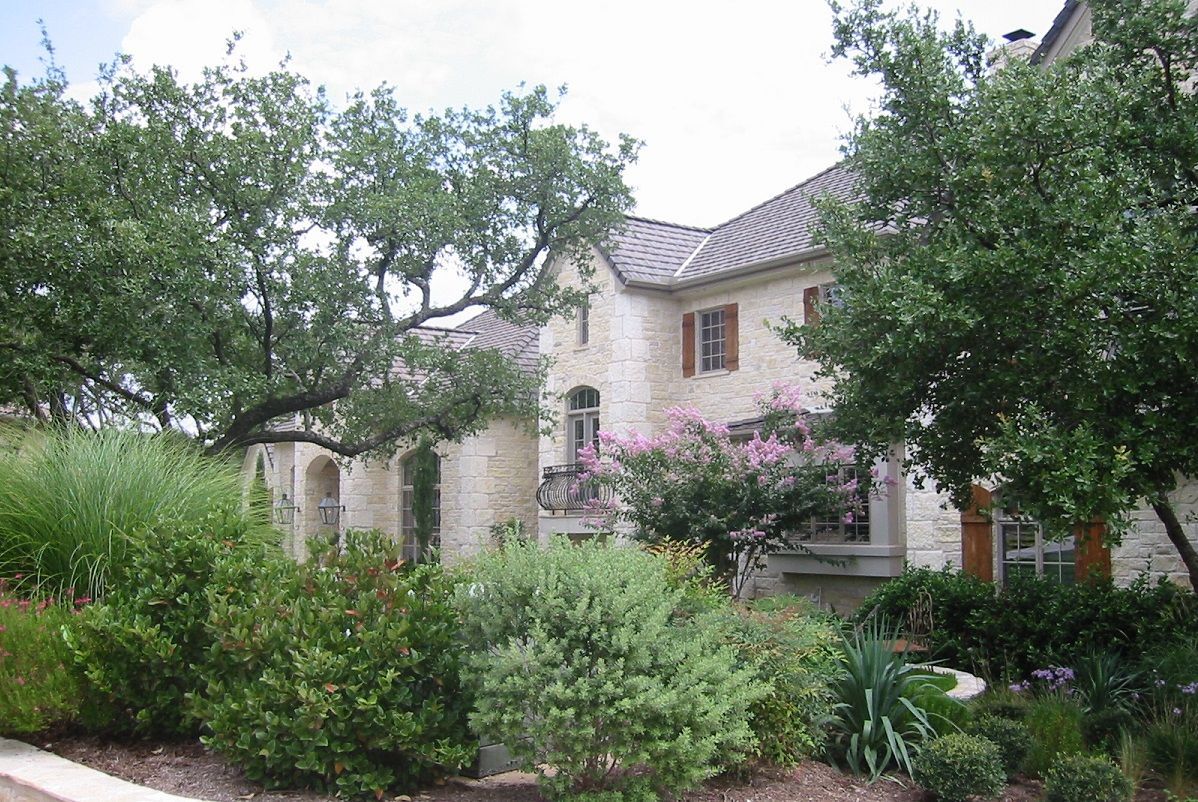 Stone house with brown shutters, nestled among trees and bushes.