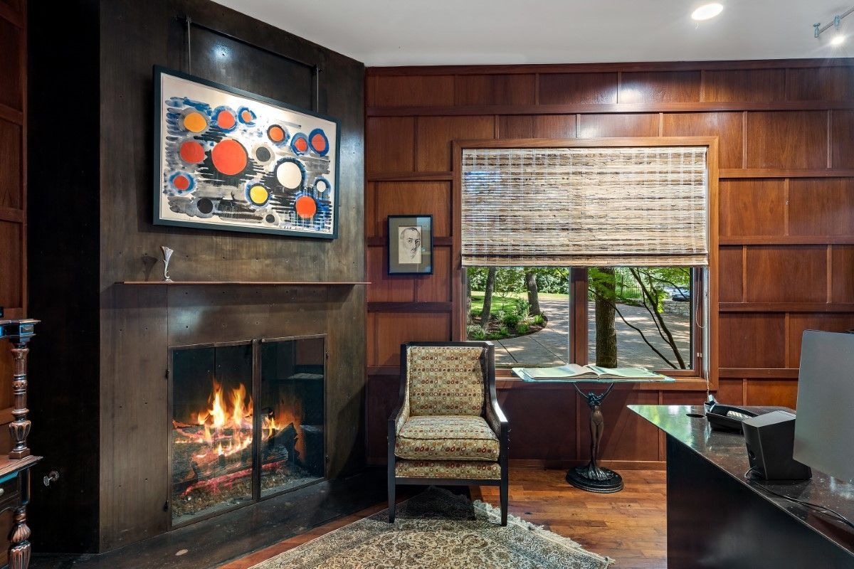 A cozy, wood-paneled study with a fireplace, art, and a window overlooking a lush yard.