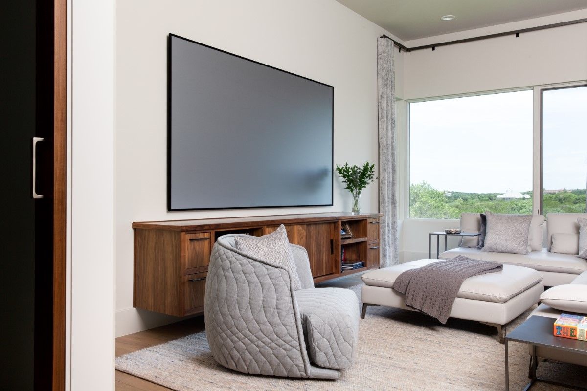 Modern living room with large TV, wooden cabinet, grey chair, sectional, and large window.
