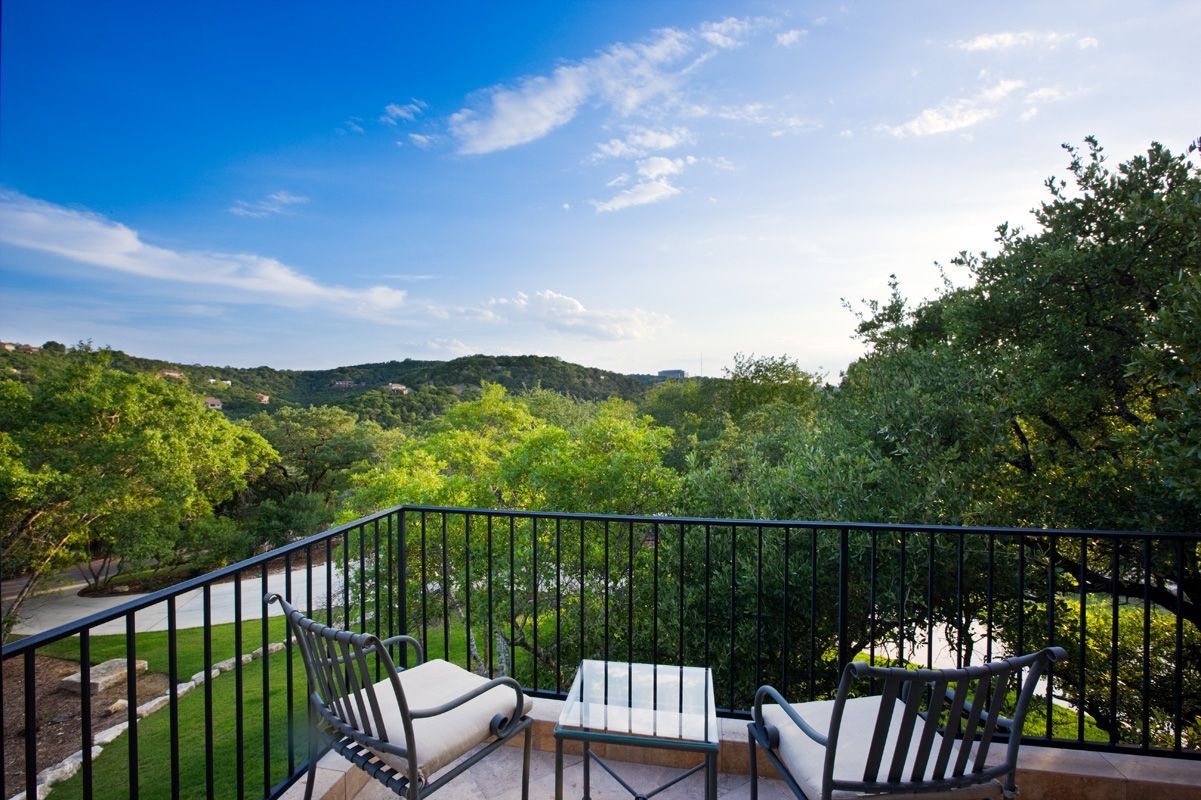 Balcony with wrought iron railing overlooking green trees under a blue sky; two chairs, small table.