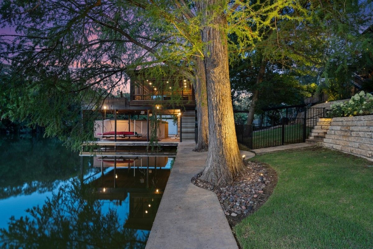 Waterfront home with dock and mature trees, reflected in calm water at dusk.