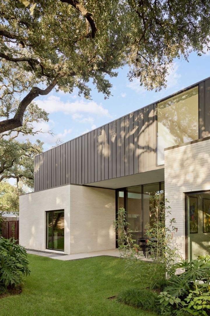 Modern home with stone walls, metal roof, and large windows, set in a green yard under a tree.