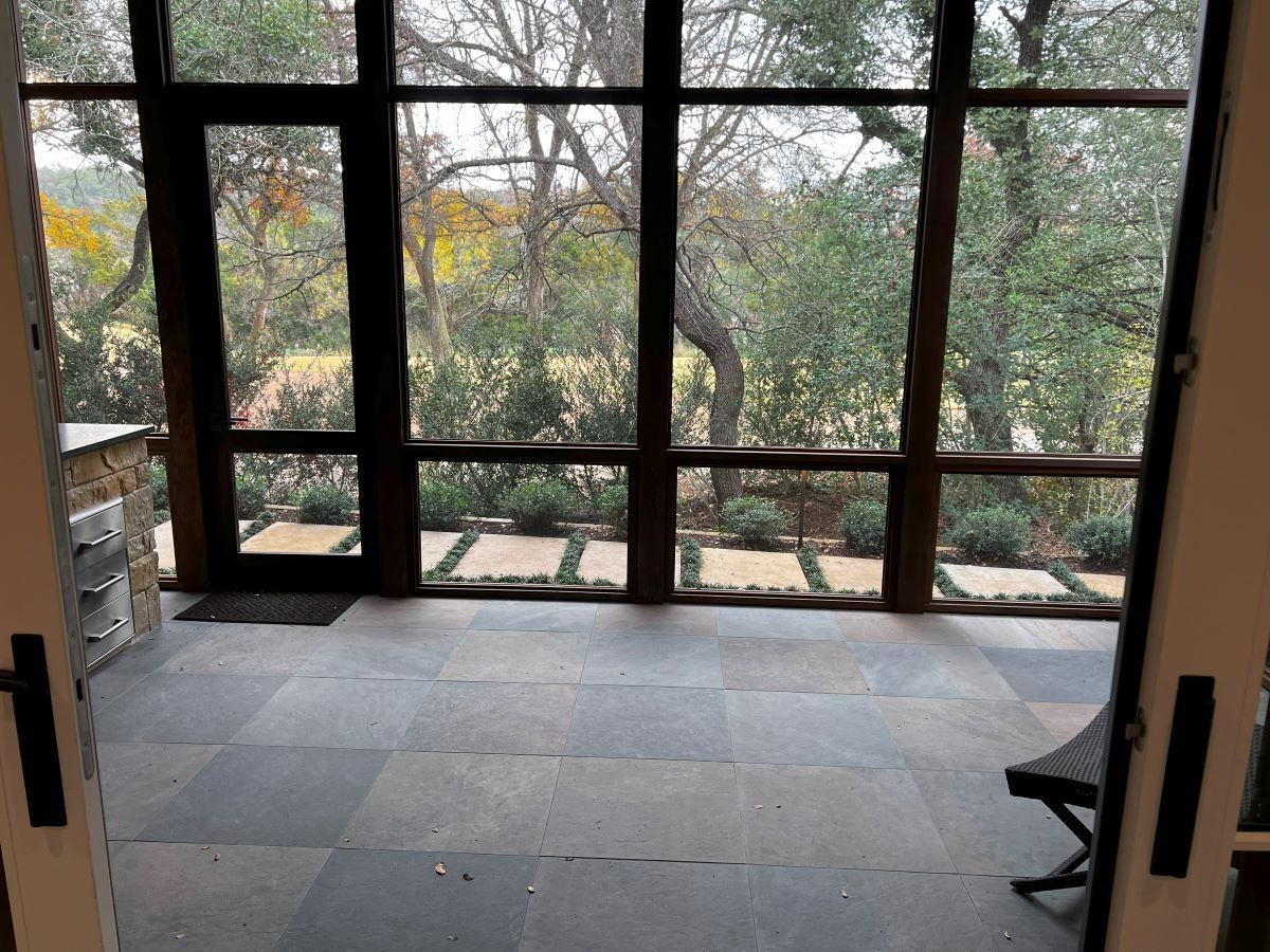 Patio with large windows overlooking a garden, gray stone floor, dark-framed windows and a black chair.