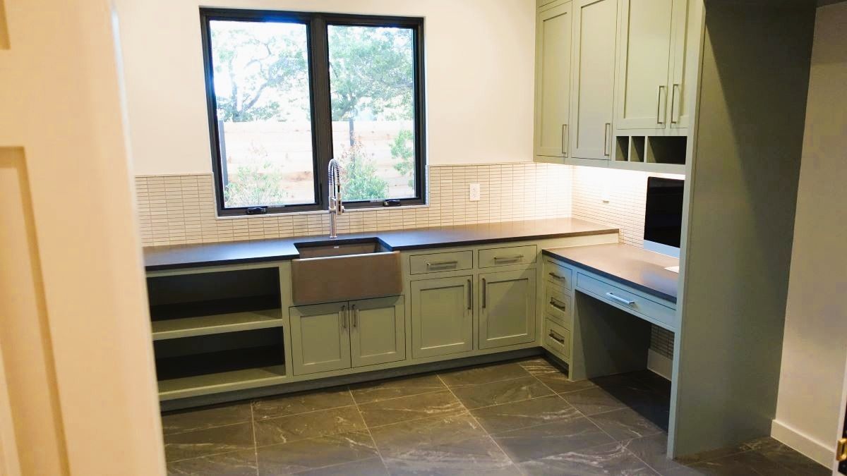 Green-painted laundry room with a sink, desk, and cabinets. Natural light streams in from a window.