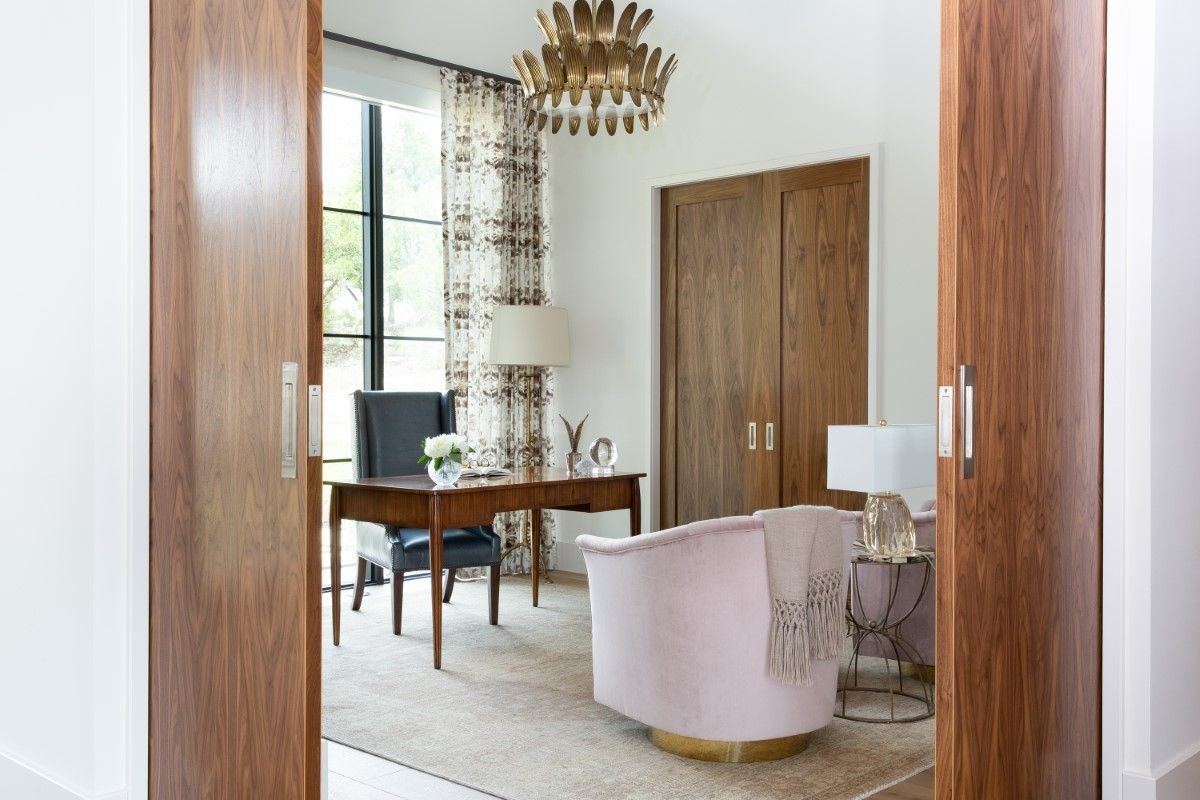 Home office with wooden doors, desk, pink chair, and gold chandelier.