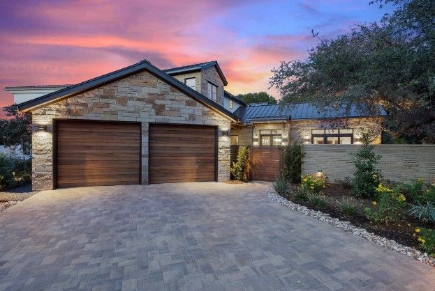 Stone house with brown garage doors and paver driveway at dusk.