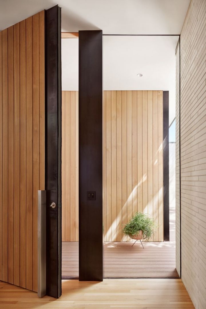 Wooden door with dark metal frame, leading to a light-filled entryway with wood paneling and a small potted plant.