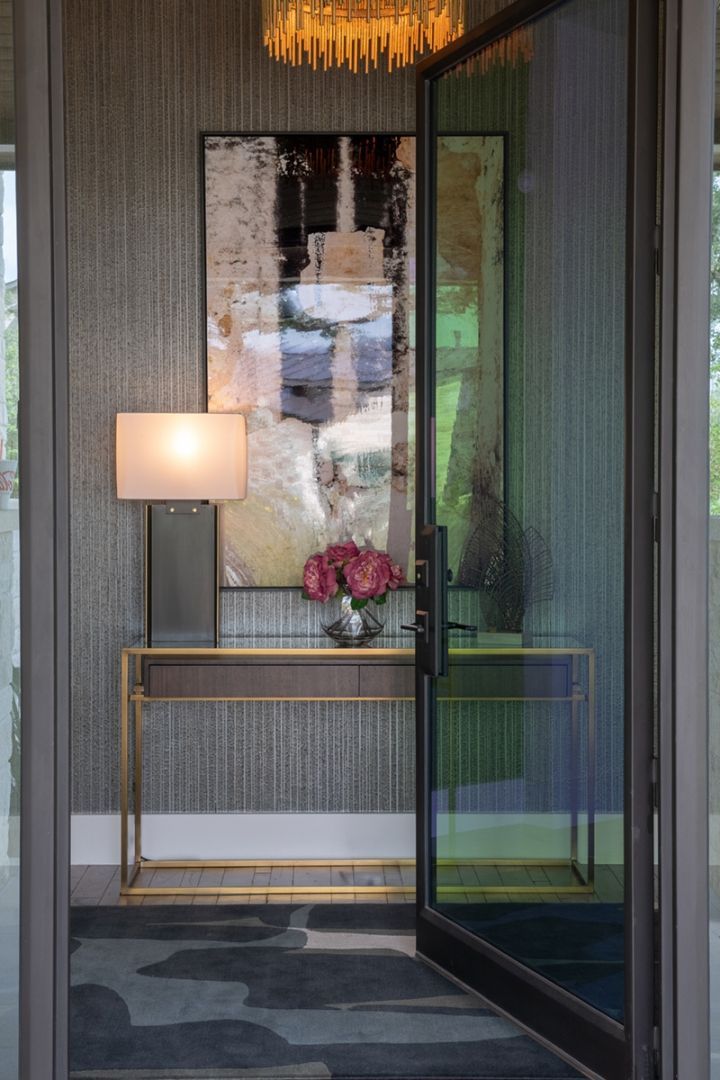 Entryway with console table, lamp, artwork, flowers, and open glass door. Gold and gray tones.