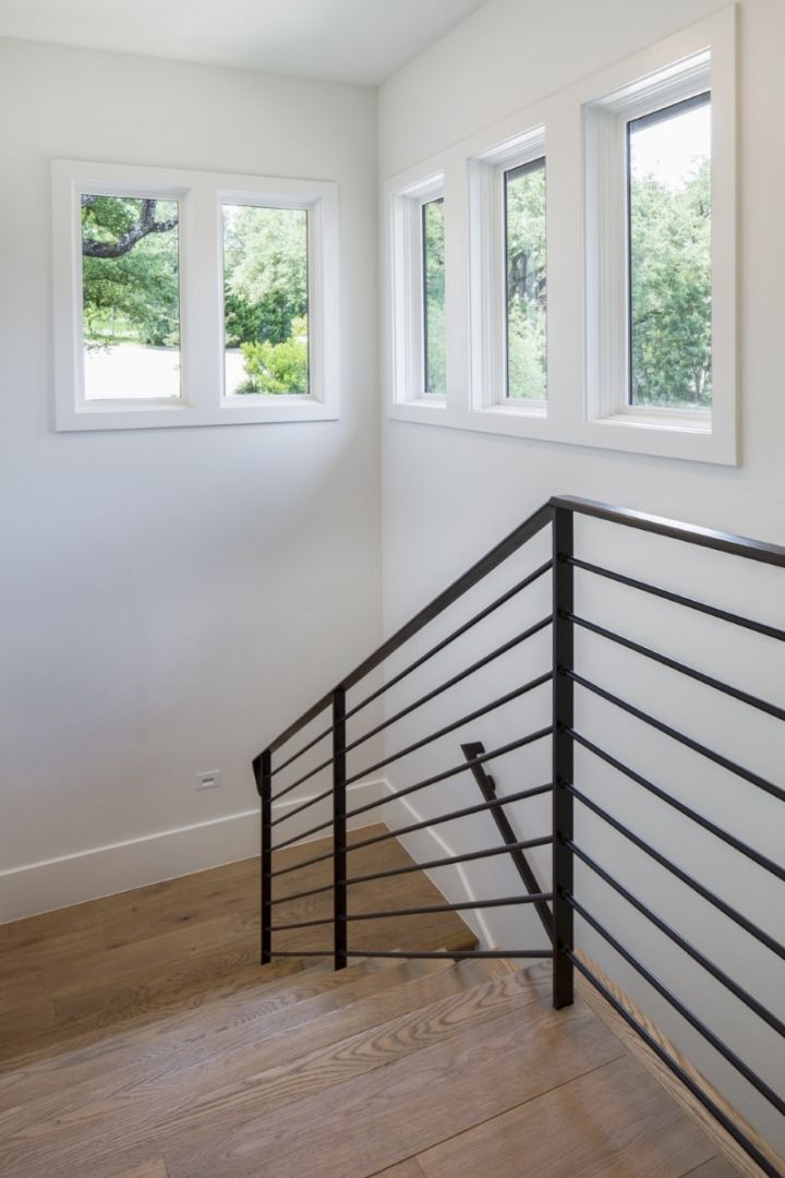 Staircase with black metal railing, light wood floor, and white walls with windows overlooking green trees.