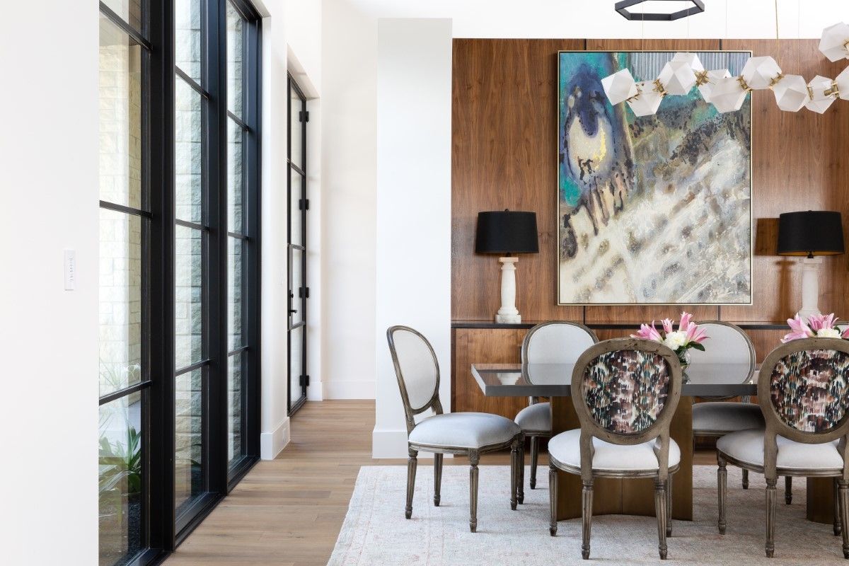 Dining room with wood-paneled wall, floral chairs, and large artwork. Black framed windows on the left.
