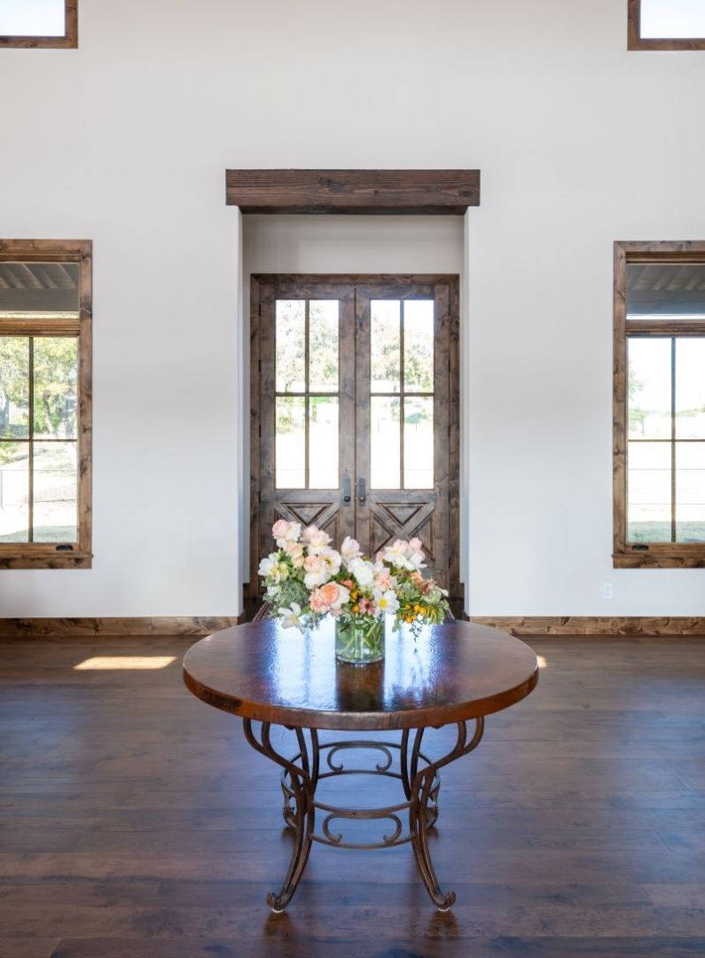 Round wooden table with floral arrangement in front of rustic double doors and windows.