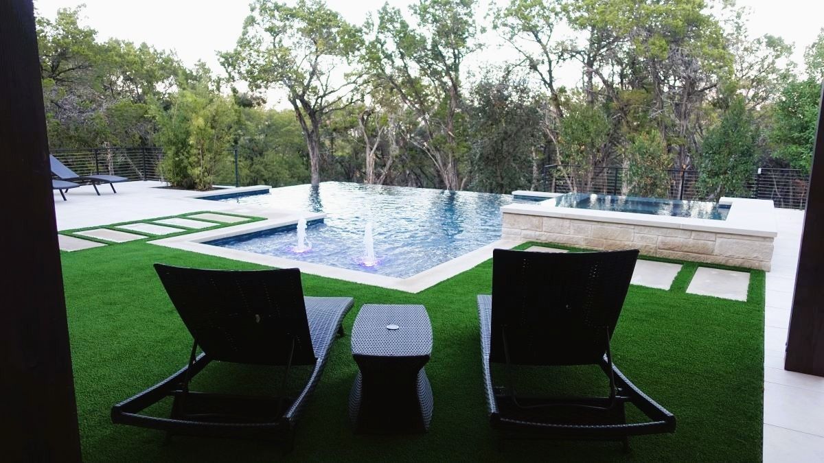 Two lounge chairs and a small table on green lawn, overlooking a pool and trees.