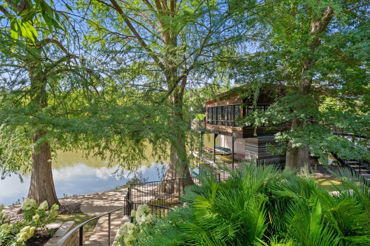 Lakeside house nestled among large trees, overlooking calm water. Lush green foliage and a pathway.