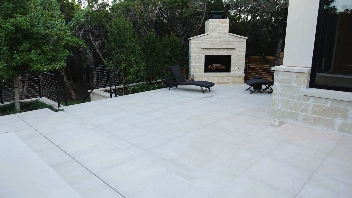 Light stone patio with fireplace, lounge chair, and trees.