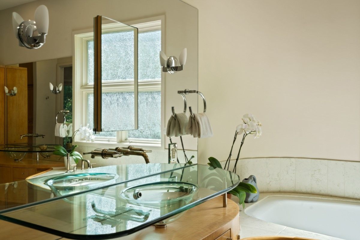 Modern bathroom with glass vanity and sinks, window with light streaming through, and a bathtub.