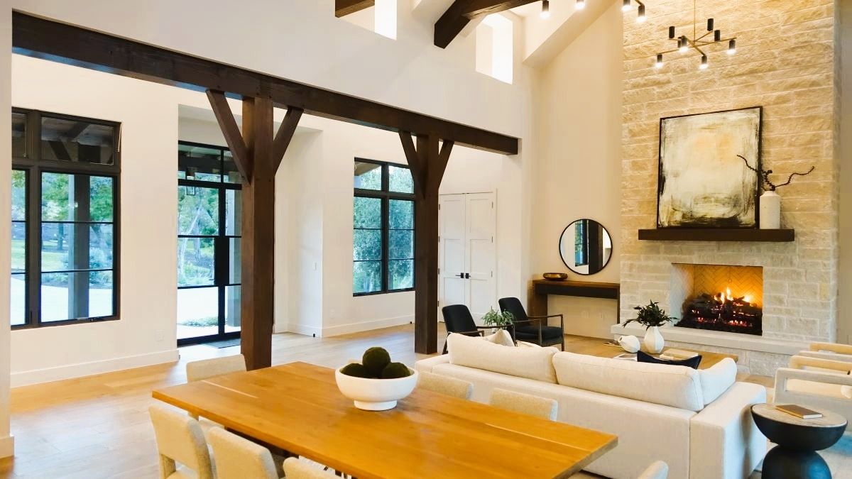 Spacious living room with a fireplace, dining table, and large windows. Light wood beams and white walls.