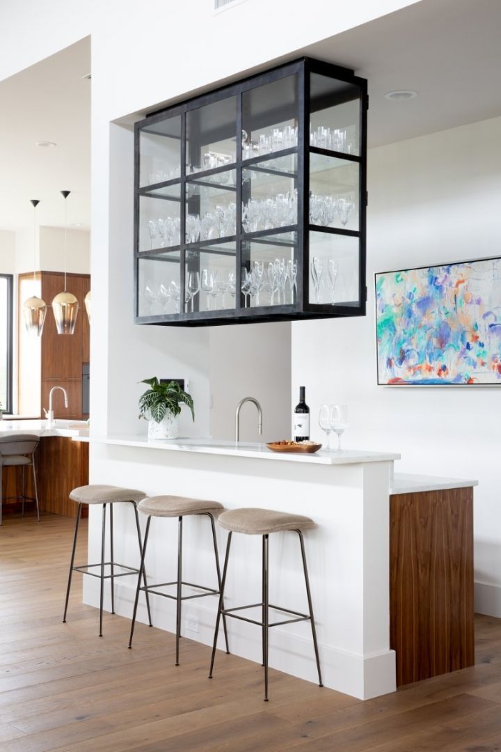 A modern bar area with a glass cabinet, stools, and artwork. Light-colored wood floors and white walls.