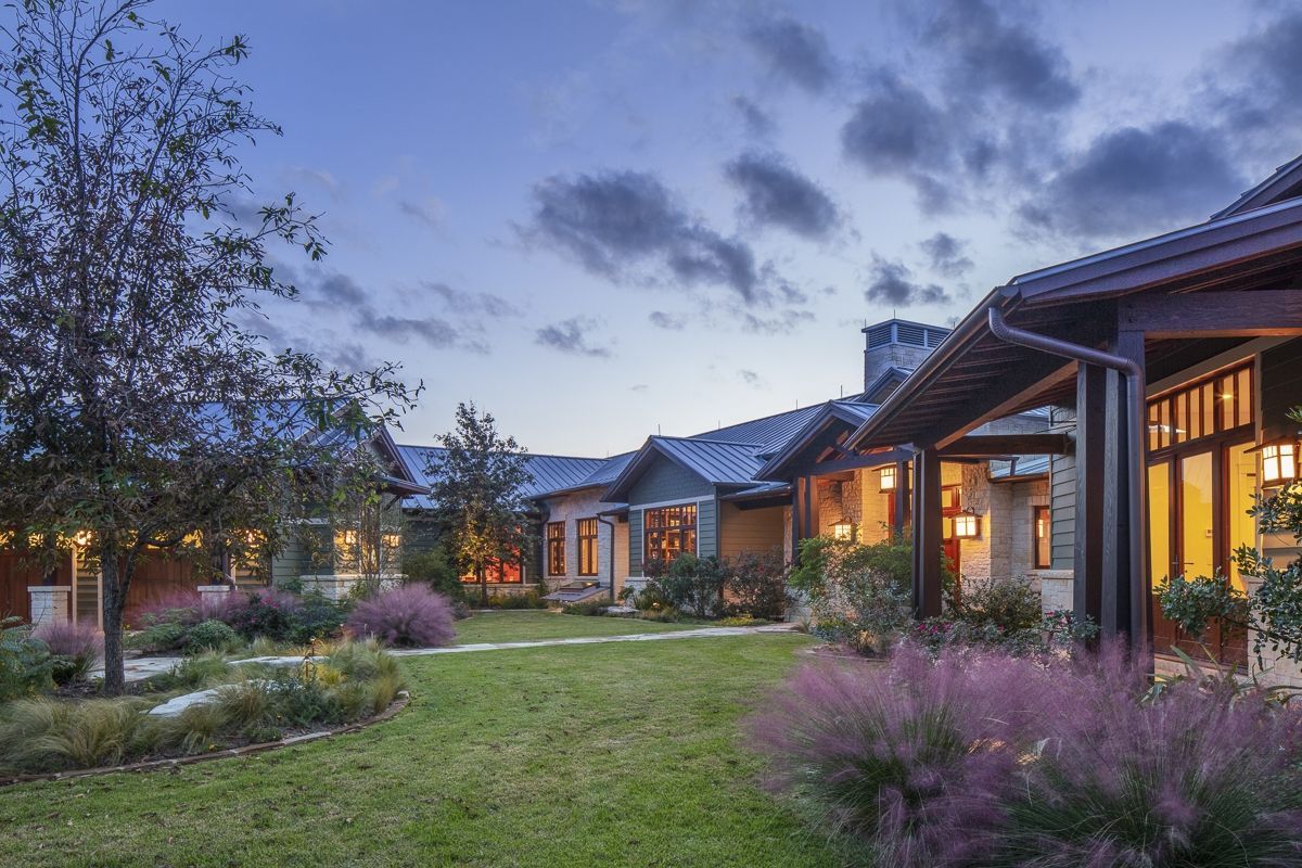 Lush lawn and garden with stone house and porch glowing with interior lights at dusk.