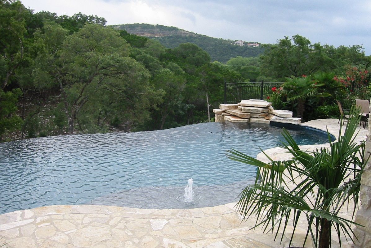 Infinity pool overlooking a forested landscape. Blue water, beige stone, green trees.