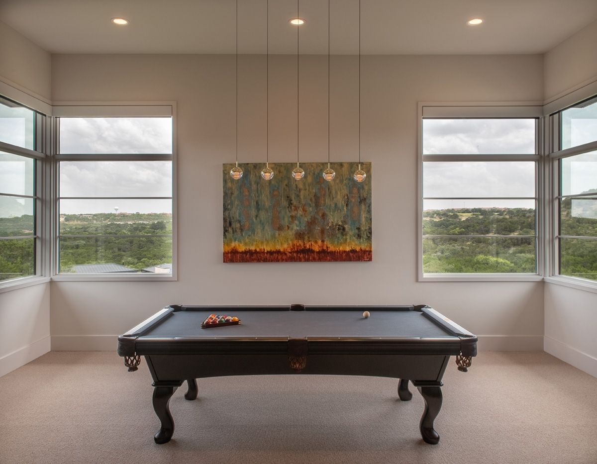 A pool table centered in a room with large windows, artwork, and three pendant lights.
