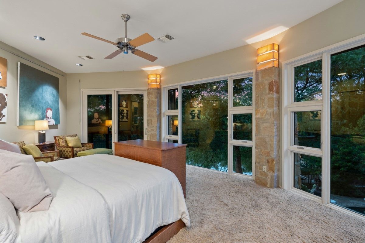 Bedroom with large windows, bed, seating area, and ceiling fan. Beige walls and carpet, natural light.
