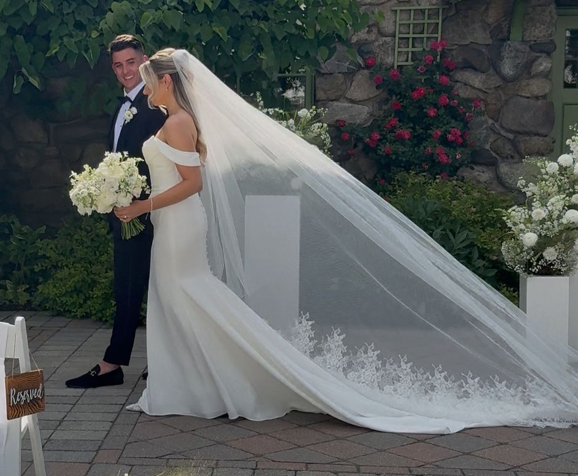 A couple walking outdoors at a wedding, with the bride wearing a long, lace-trimmed veil and holding white flowers.