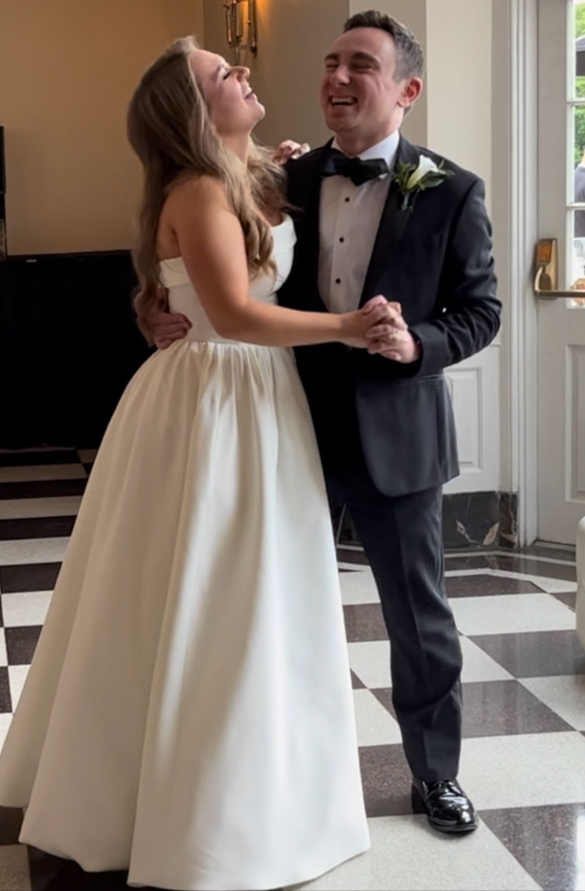 A laughing couple in wedding attire dances together on a checkered floor in a bright, sunlit room.