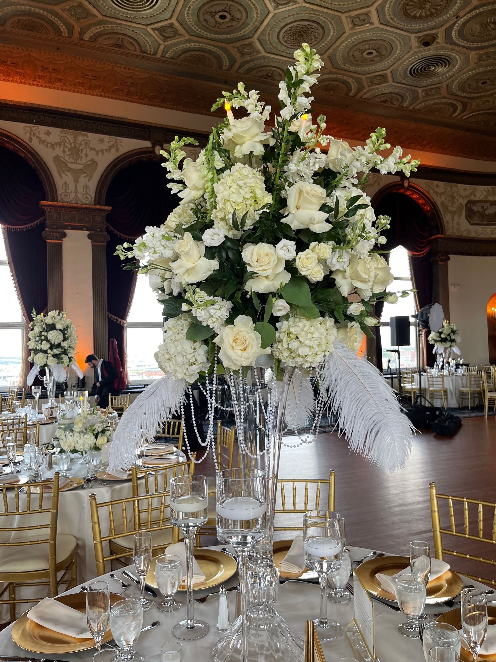 Tall, crystal vase centerpiece with white floral arrangements and feathered accents in an elegant ballroom setting.