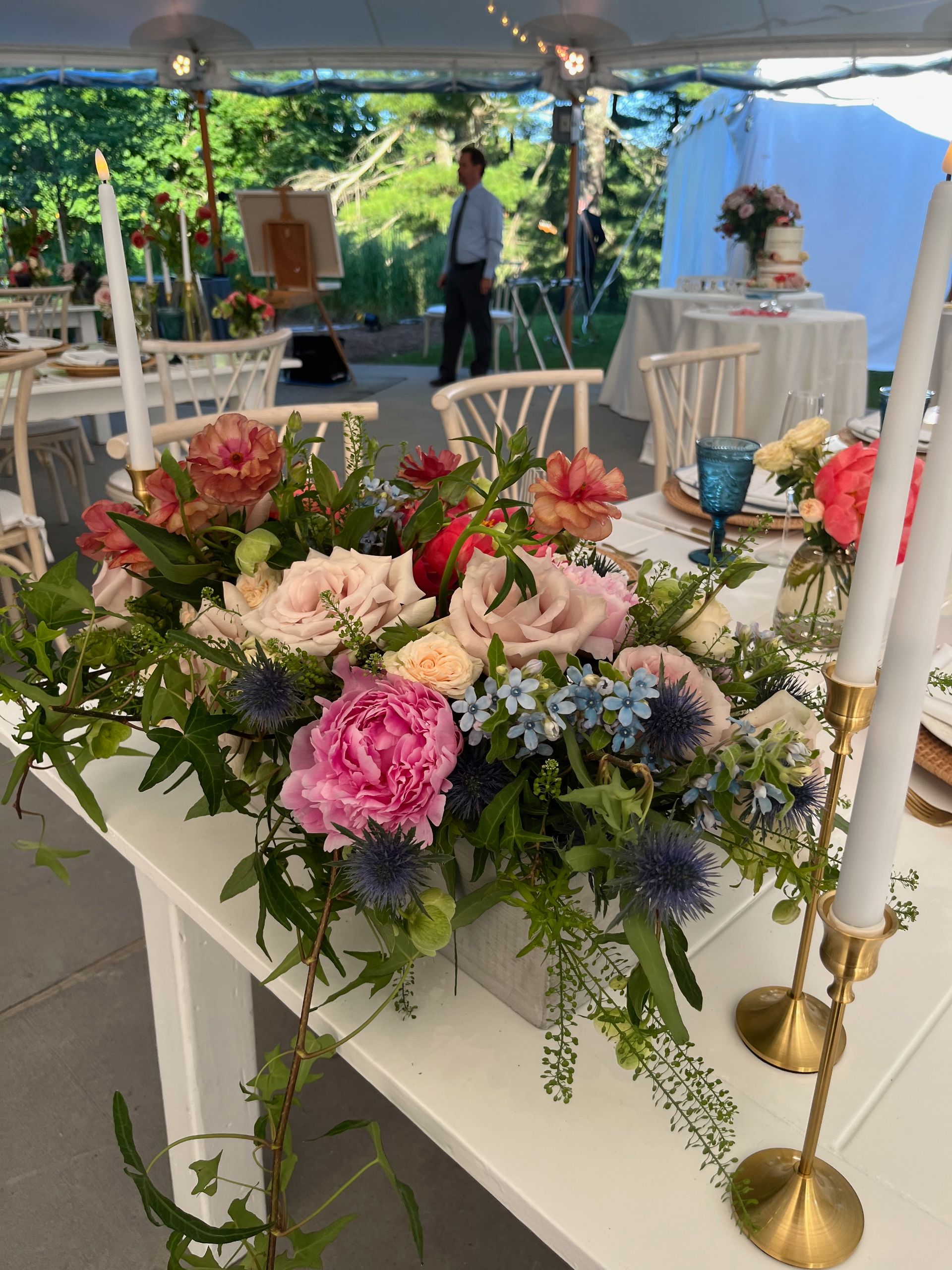 An outdoor wedding reception table featuring a floral centerpiece with pink, peach, and blue flowers near gold candlesticks.