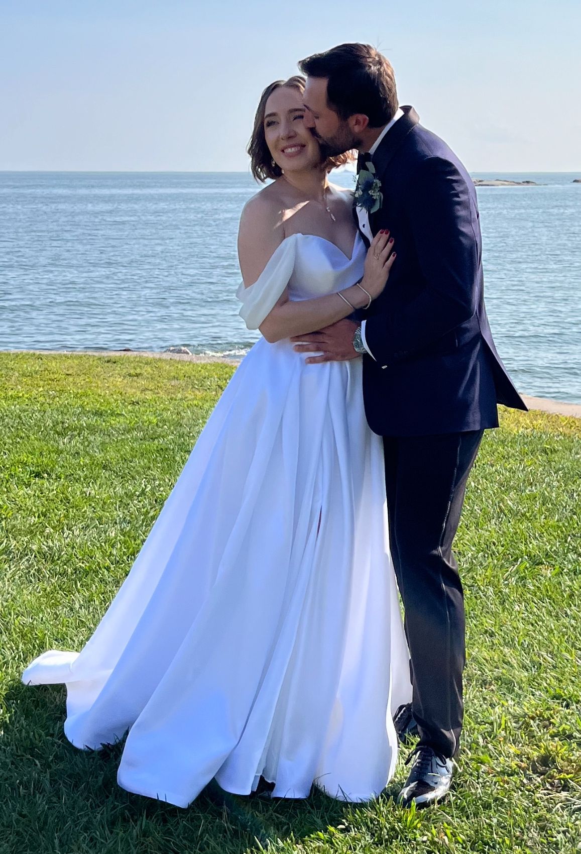 A bride in a white wedding gown and a groom in a dark suit embracing and kissing by the ocean on a sunny day.