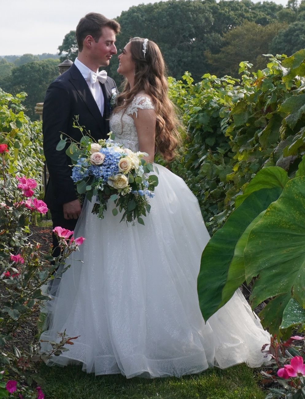 A couple faces each other in a wedding garden, the bride holding a bouquet of blue and white flowers.