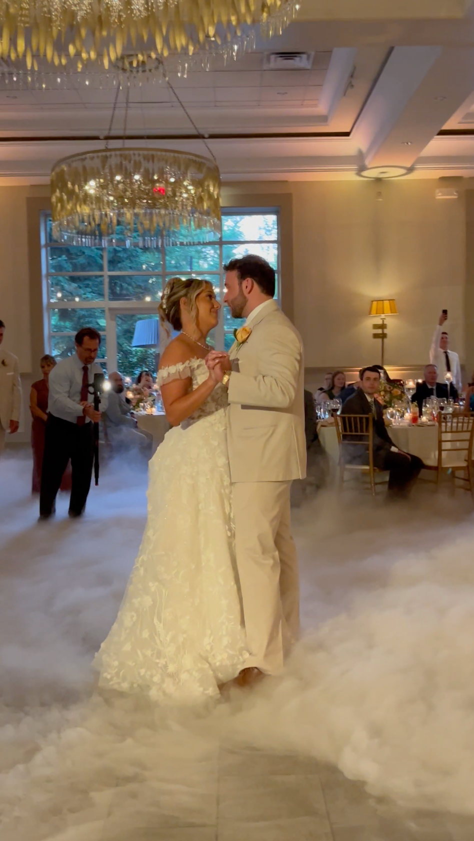A couple in wedding attire shares a first dance on a dance floor covered in low-lying fog in an elegant banquet hall.