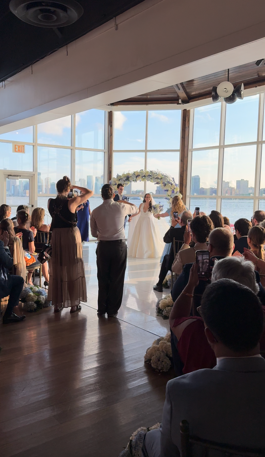 A bride walks down the aisle during an indoor wedding ceremony with large windows overlooking a city skyline.