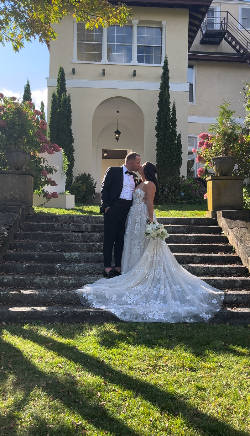 A bride and groom kiss on stone steps in front of a historic building with white walls and tall, thin trees.