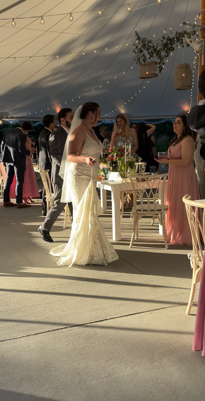 A bride in a white gown walks through a tented wedding reception as guests mingle around tables under string lights.