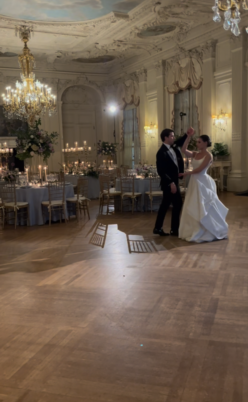 A couple in formal attire dances in a ballroom with ornate ceilings, chandeliers, and round dinner tables.