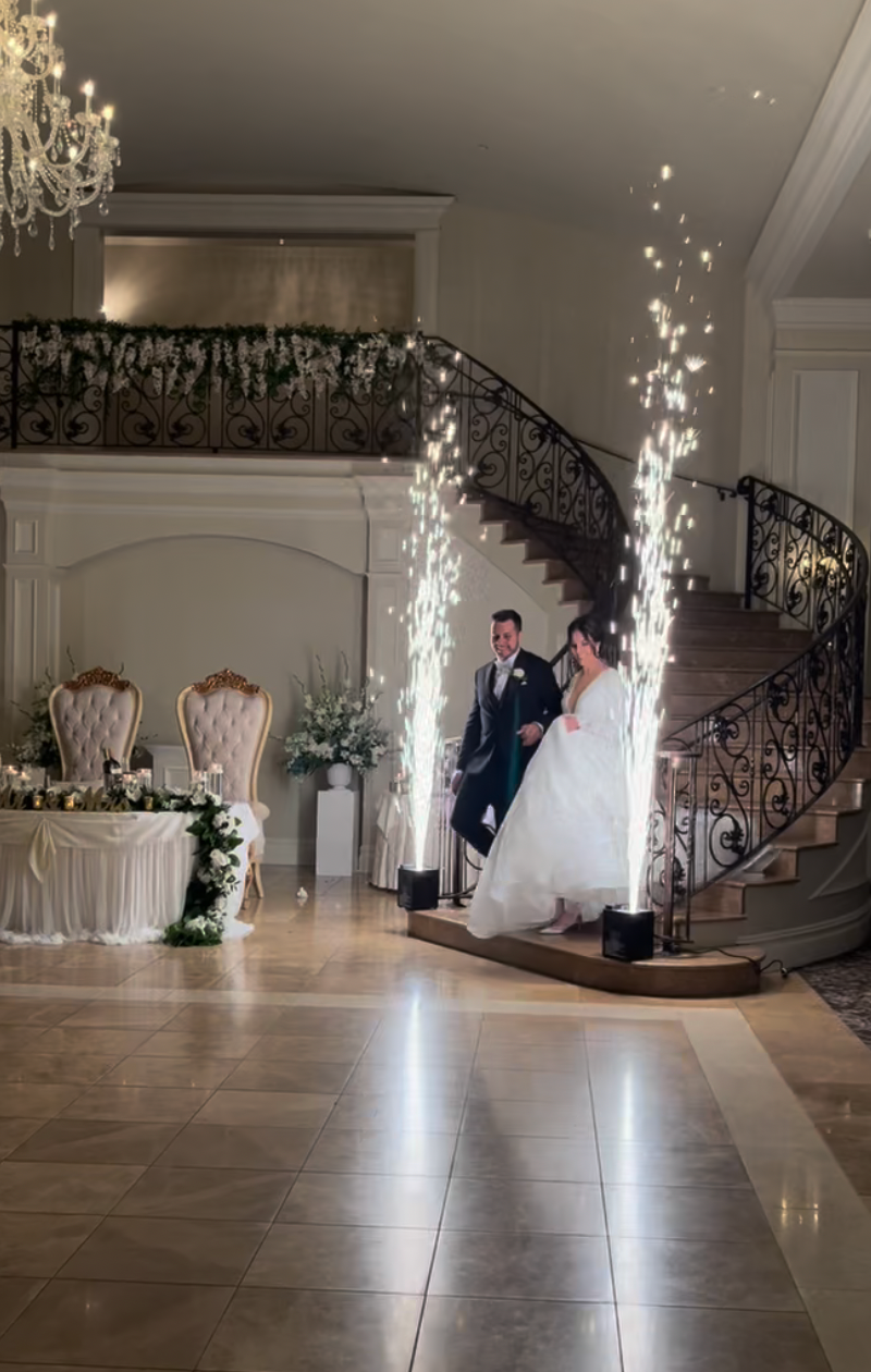 A bride and groom walk down a grand staircase at a wedding reception, flanked by two vertical sparkler fountains.