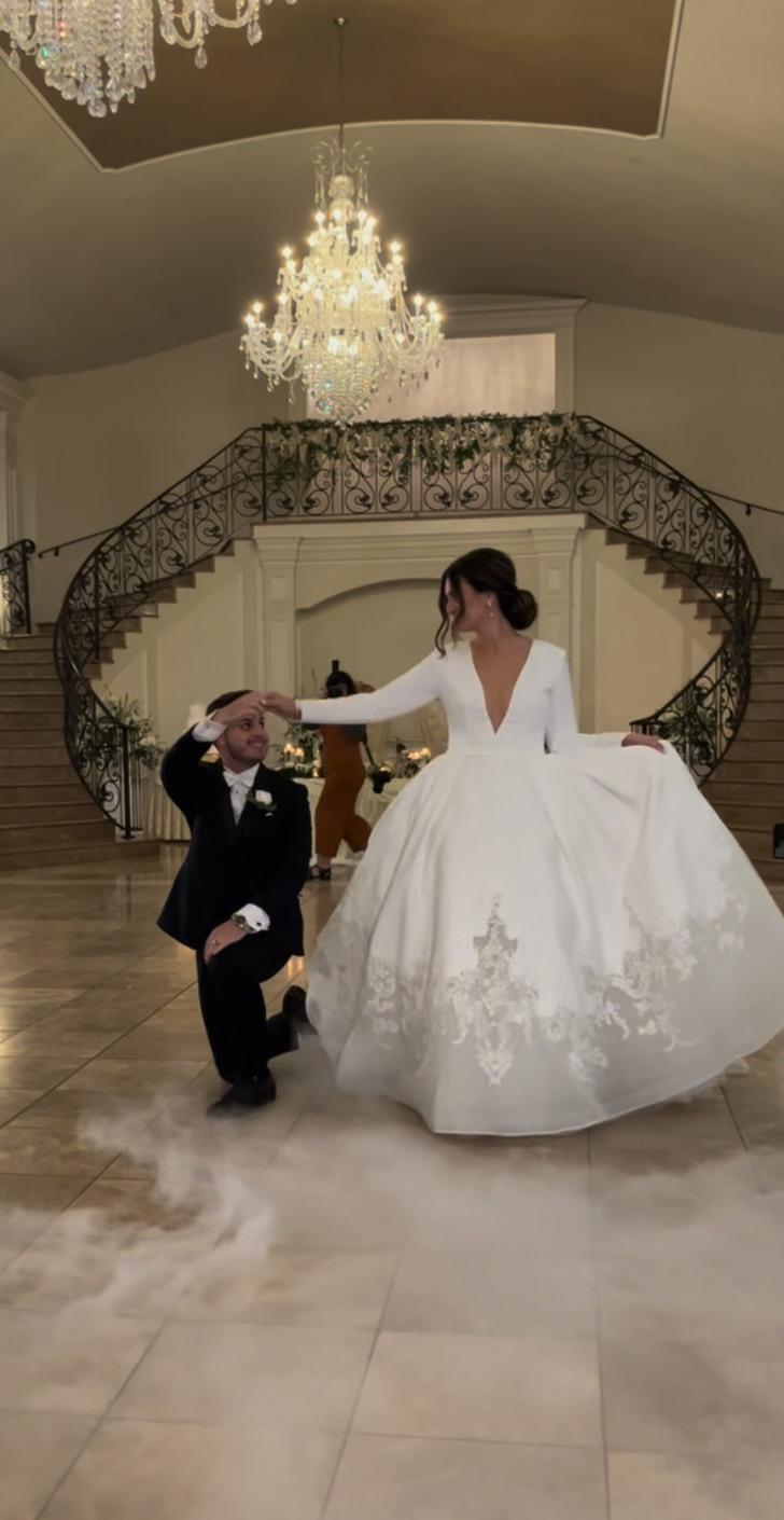 A bride in a full white gown dances with a groom in a black suit inside a grand hall with a chandelier and staircase.