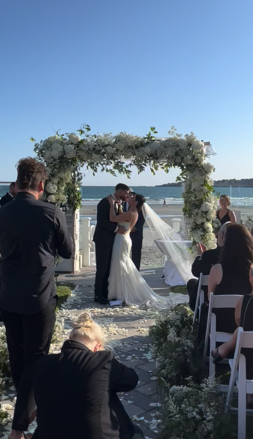 A bride and groom kiss under a large floral arch on a sandy beach during their wedding ceremony.