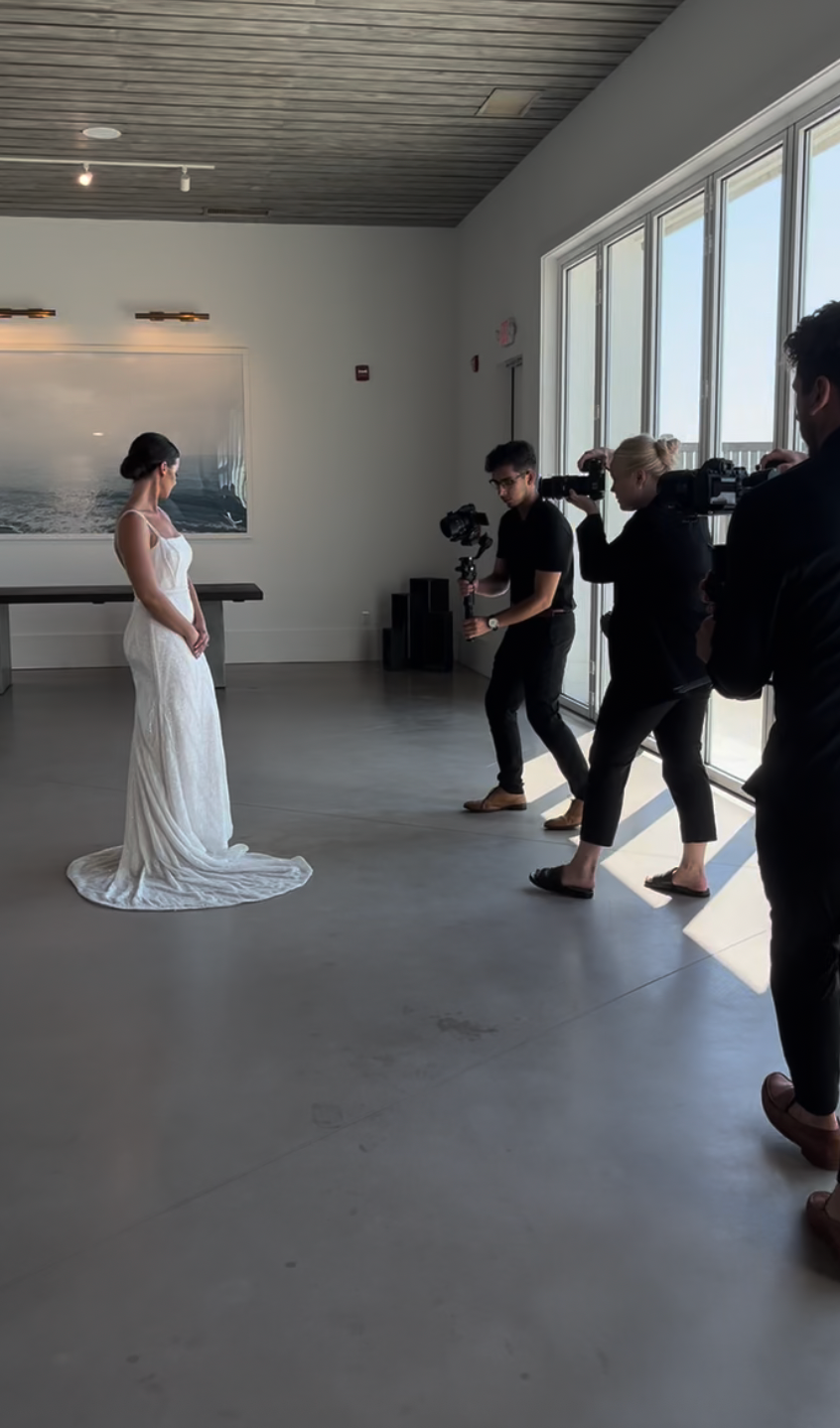A bride in a white wedding gown stands in a bright room while being filmed by two videographers near a window.